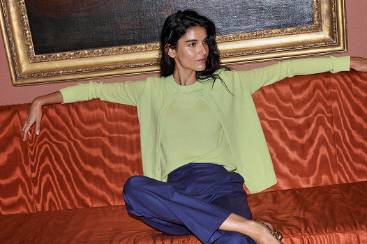 A woman sits on a rust-orange patterned sofa wearing a light lime green long-sleeve top and dark blue pants, with a large gold-framed painting behind her.