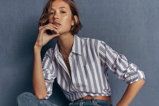 A woman wearing a white button-down shirt with thin blue vertical stripes, tucked into blue jeans with a brown belt, posed against a textured slate blue background.