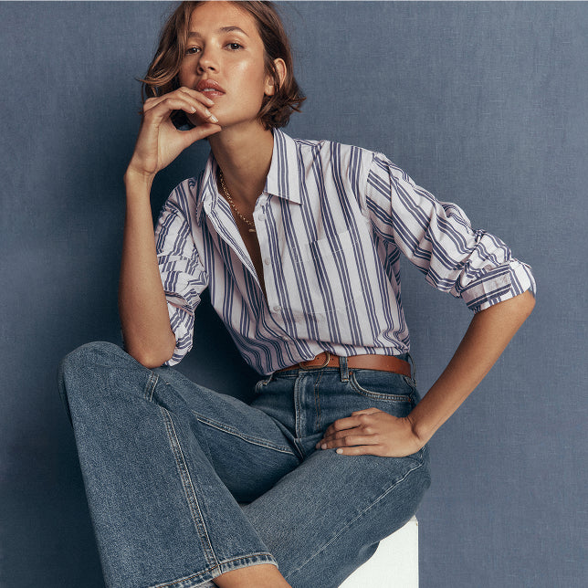 A woman seated against a textured blue-gray wall wearing a white button-up shirt with thin blue vertical stripes, tucked into blue jeans with a brown belt.