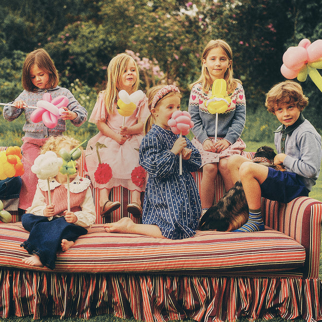 Six children sitting on a striped daybed in an outdoor garden setting, each holding a colorful balloon shaped like an animal, with a dog lying beside them.