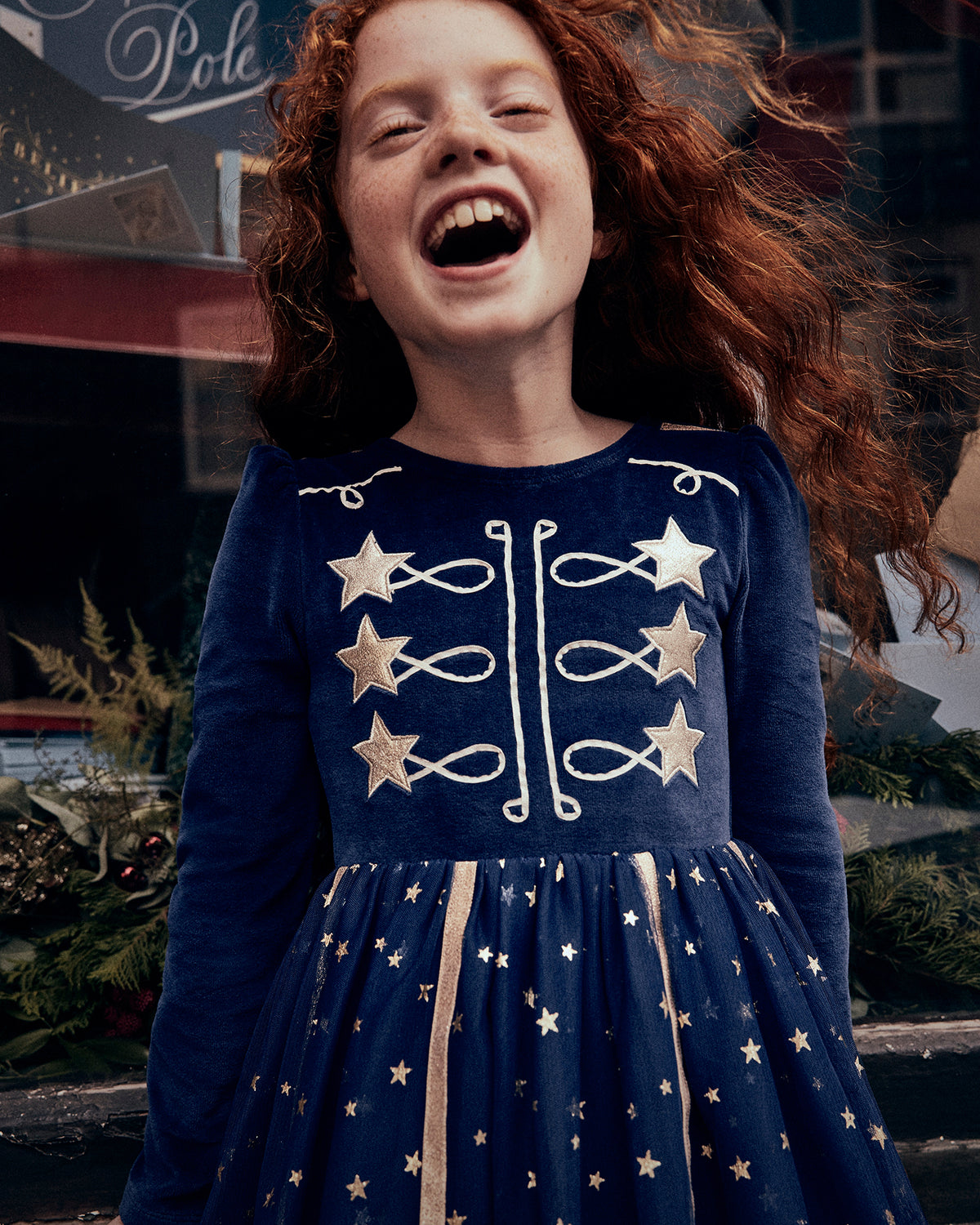 A young girl with long red hair wearing a navy blue dress with gold star and bow details, captured in a joyful moment with her eyes closed and mouth open, set against a festive outdoor background.
