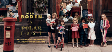 A group of children and two women stand in front of a shop window decorated for Christmas, with large overlaid text reading 'MINI BODEN x POLAR POST' and smaller text below. A red British postage box is visible on the left.