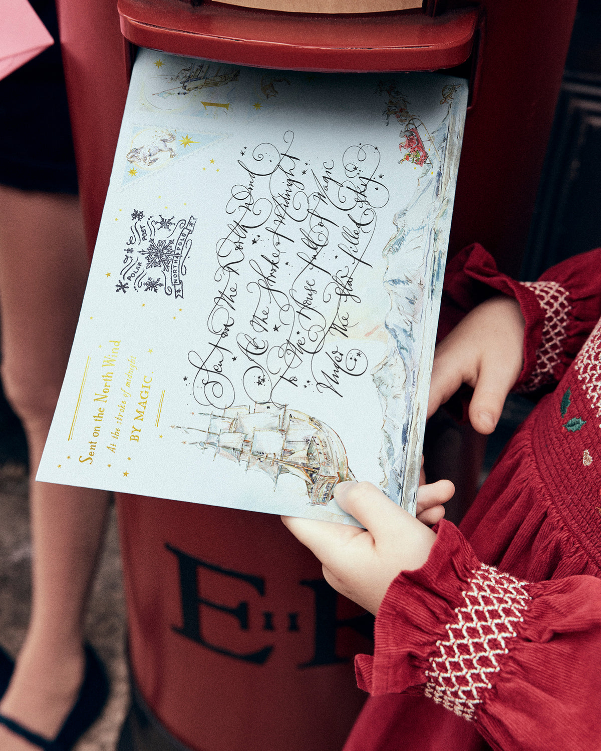 A child in a red dress holds a printed card or booklet with decorative script and a ship illustration, standing beside a red post box.