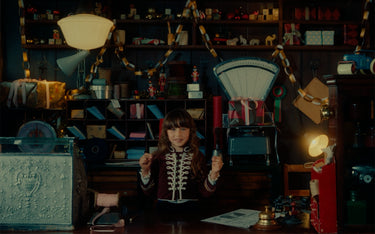 A young girl with dark hair sits at a desk in a dimly lit, cluttered vintage-themed room filled with toys, shelves, and warm lamps. She is wearing a dark red and white embroidered jacket and holding two small toy cars.