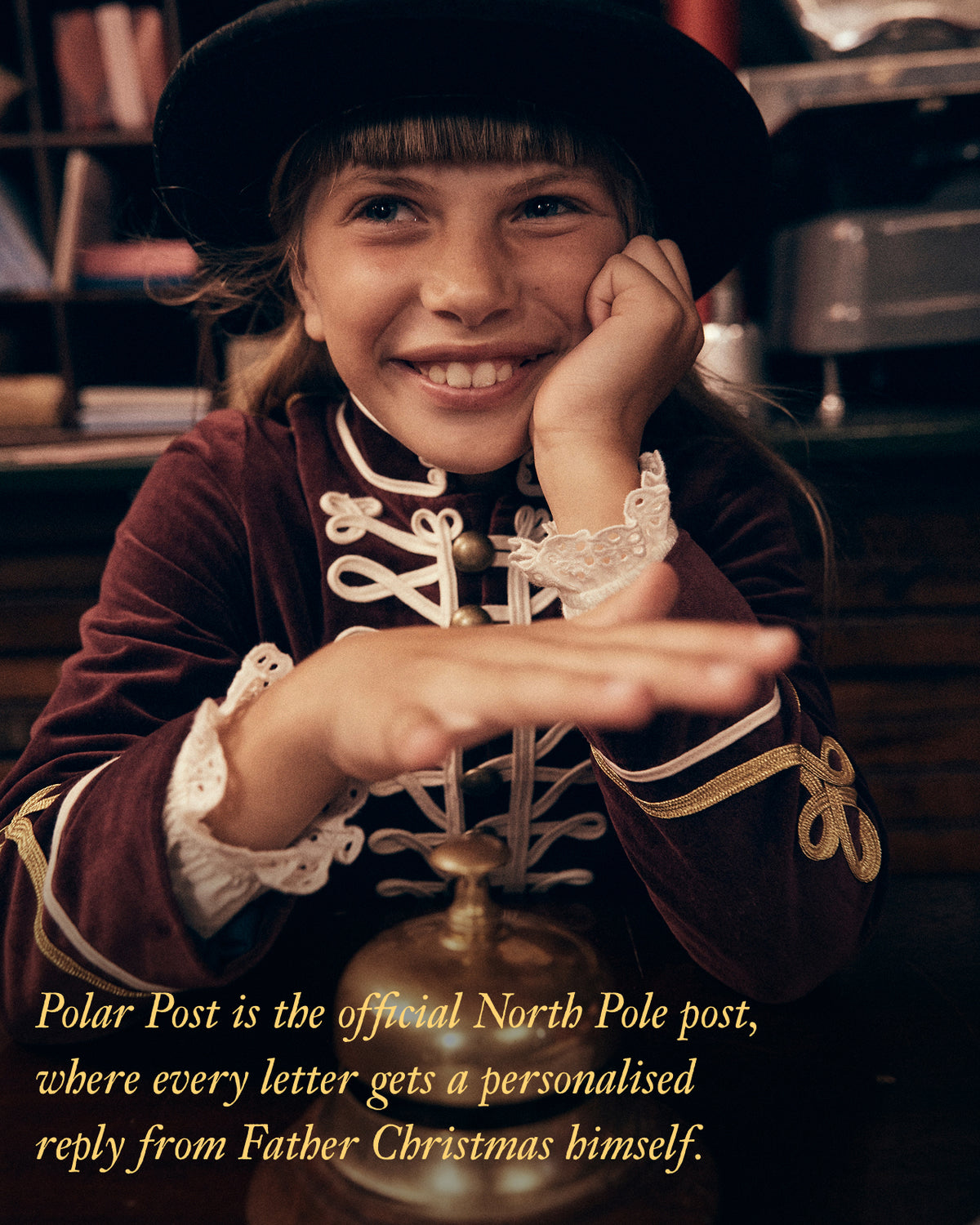 A smiling young girl in a black hat and a dark burgundy velvet Christmas-themed costume with gold trim and white lace cuffs poses indoors. A brass bell is in the foreground, and text at the bottom reads, 'Polar Post is the official North Pole post, where every letter gets a personalised reply from Father Christmas himself.