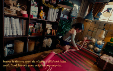 A woman kneels on the floor in a rustic, book-filled room, wrapping a pink striped fabric or ribbon around a large gift box. The scene is warmly lit with hanging lamps and shelves of books and decorative items in the background.