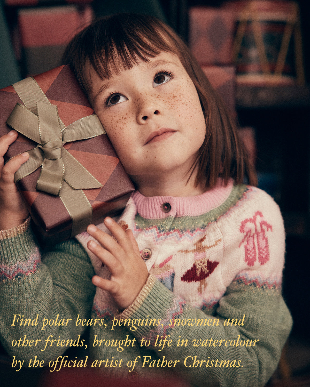 A young child with freckles holds a wrapped gift box with a large ribbon bow, wearing a knitted sweater in a warm indoor setting. Gold text at the bottom reads, 'Find polar bears, penguins, snowmen and other friends, brought to life in watercolour by the official artist of Father Christmas.