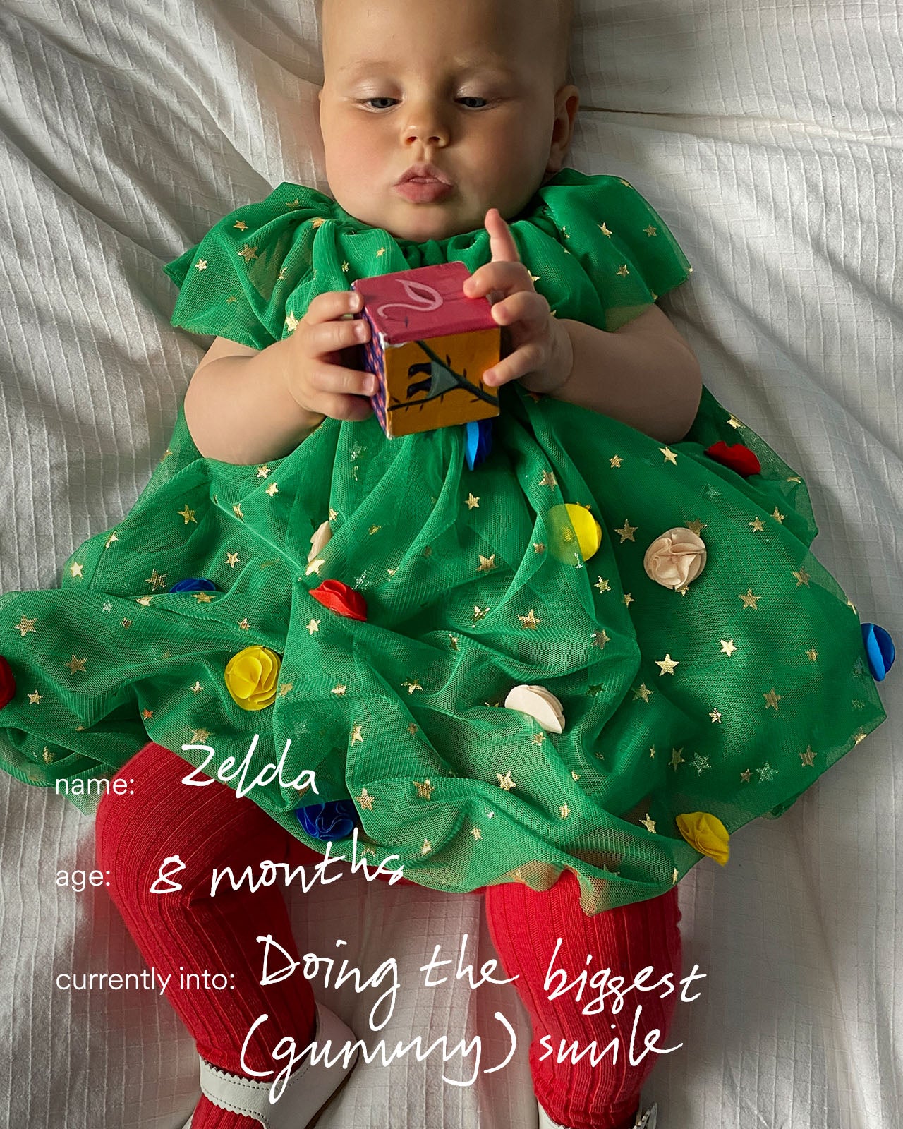 Baby in a green dress with Christmas tree design holding a toy, lying on a white blanket.