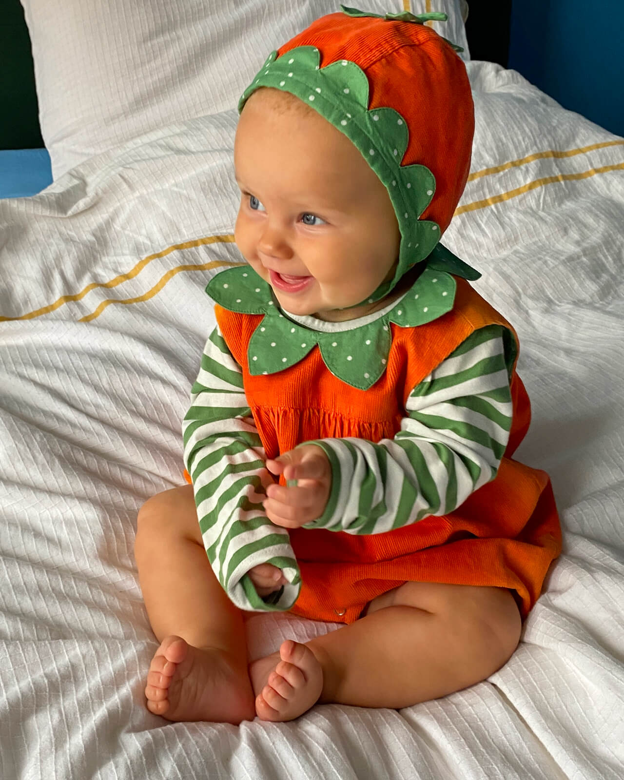 Baby wearing an orange and green outfit with a matching hat on a white bed.