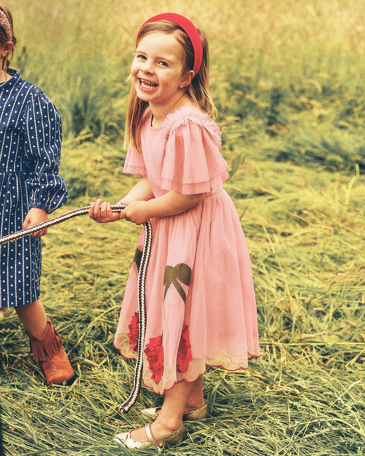 A young girl stands in a grassy field wearing a light pink dress with red floral accents, a red headband, and holding a rope. Another child in a blue polka-dot dress is partially visible on the left.