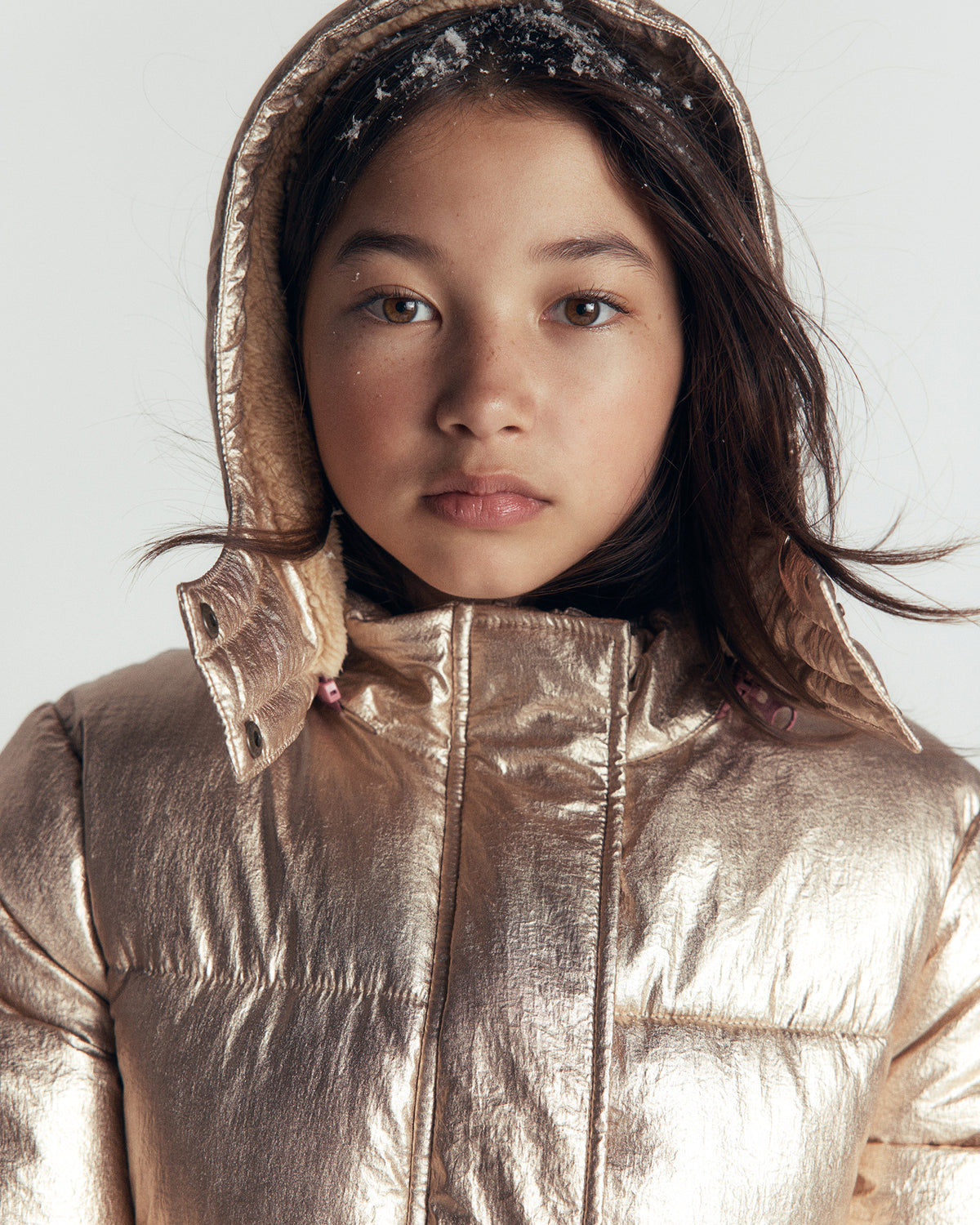 A close-up of a child wearing a shiny metallic gold puffer jacket with a hood, photographed against a plain light gray background.