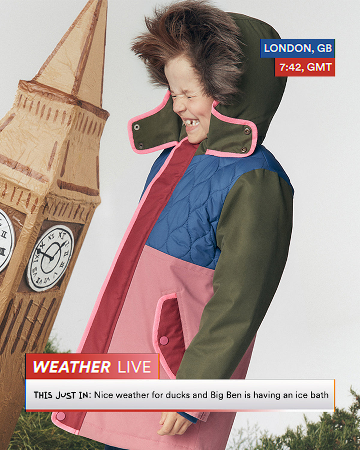 A child wearing a colorful quilted hooded jacket with pink trim, standing outdoors next to a large clock tower prop. The jacket has color-blocked sections of olive green, blue, and pink.
