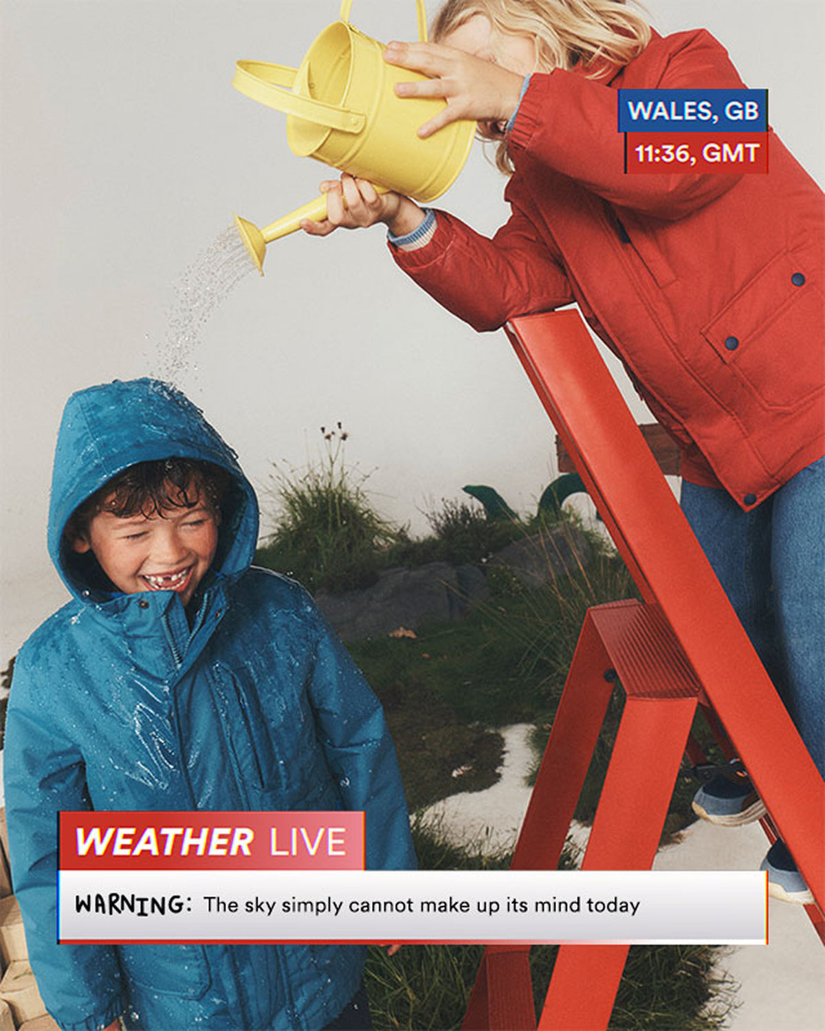 Two children wearing rain jackets play outdoors in the rain. One child stands on a red ladder pouring water from a yellow watering can while the other child stands below smiling.