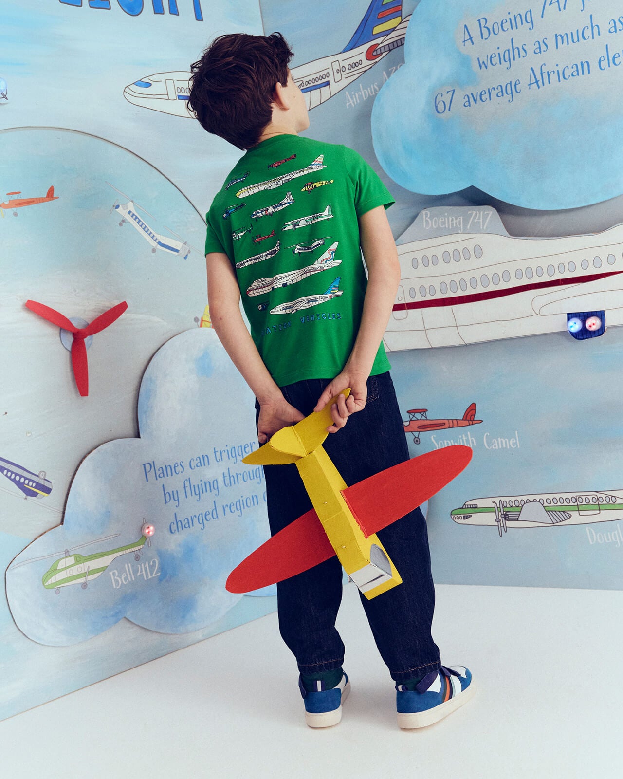 Child holding a red and yellow airplane model in front of an educational poster about airplanes.