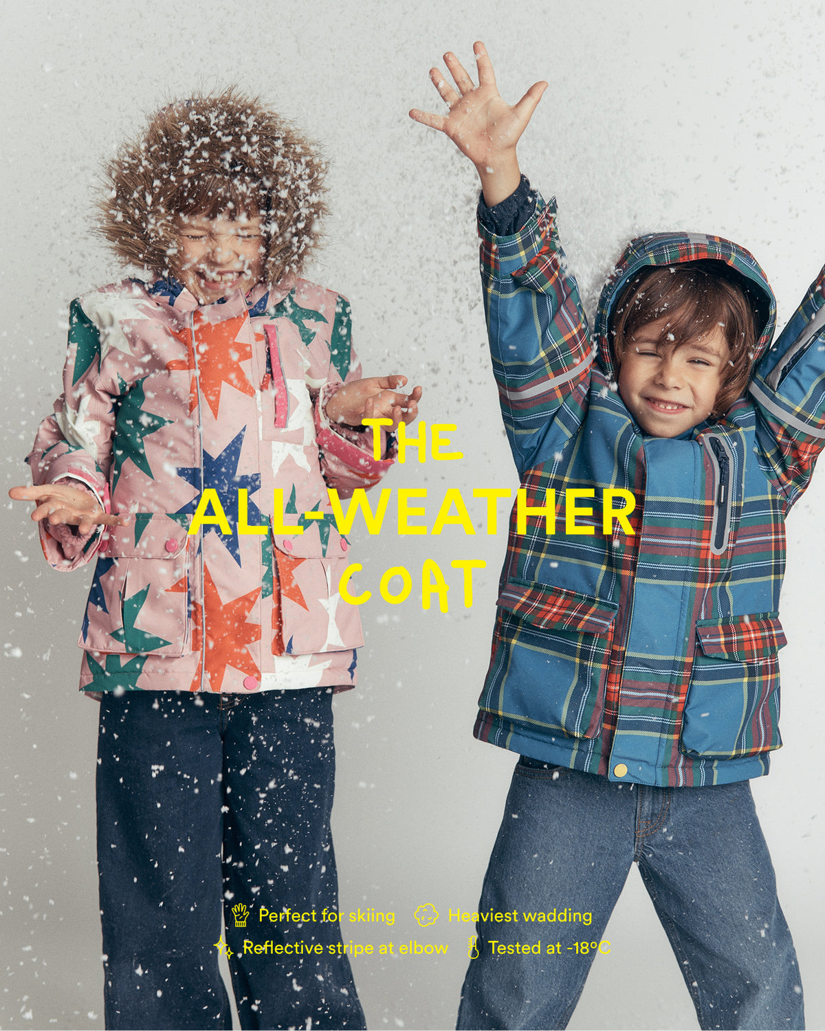 Two children wearing colorful winter jackets pose in falling snow. The child on the left wears a light pink jacket with orange and green star patterns, and the child on the right wears a blue and red plaid hooded jacket.