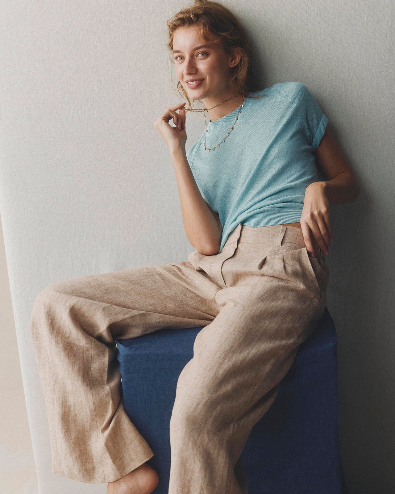 Woman wearing a light blue top and beige pants sitting on a blue stool against a plain wall.