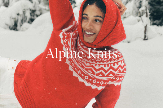 Person wearing a red knitted sweater with white patterns lying on a snowboard in the snow, with 'Alpine Knits' text overlay.