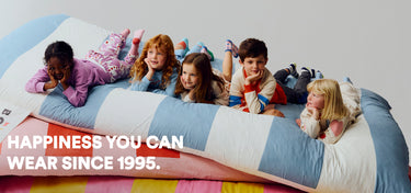 Children playing on a colourful bed with text 'Happiness you can wear since 1995'.