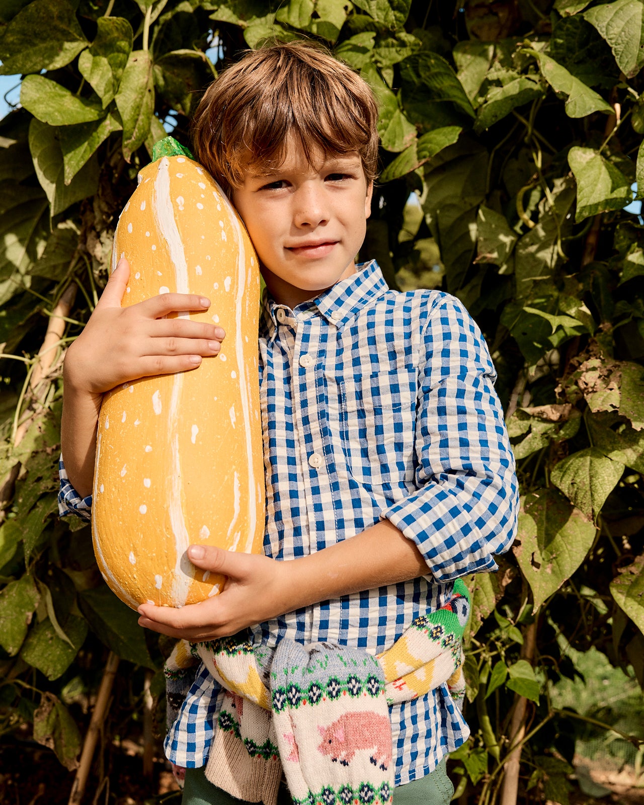 Child holding a large yellow pumpkin in an outdoor setting with greenery.