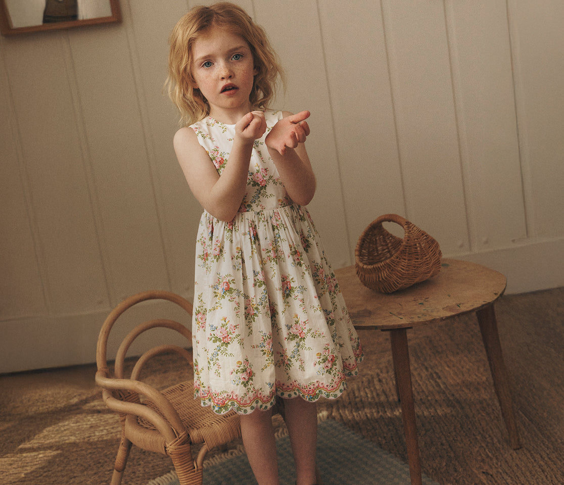 Young girl in a floral dress standing in a room with a wooden table and chair.