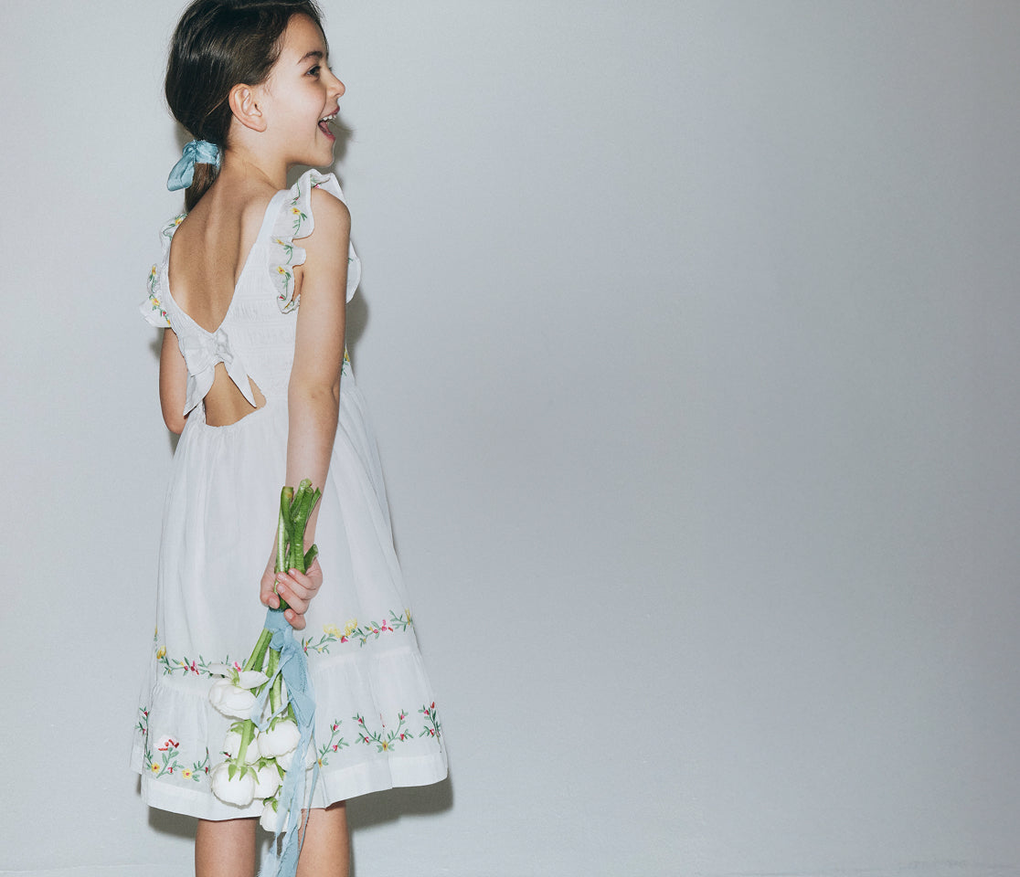 Young girl in a white dress with floral details holding flowers against a plain background