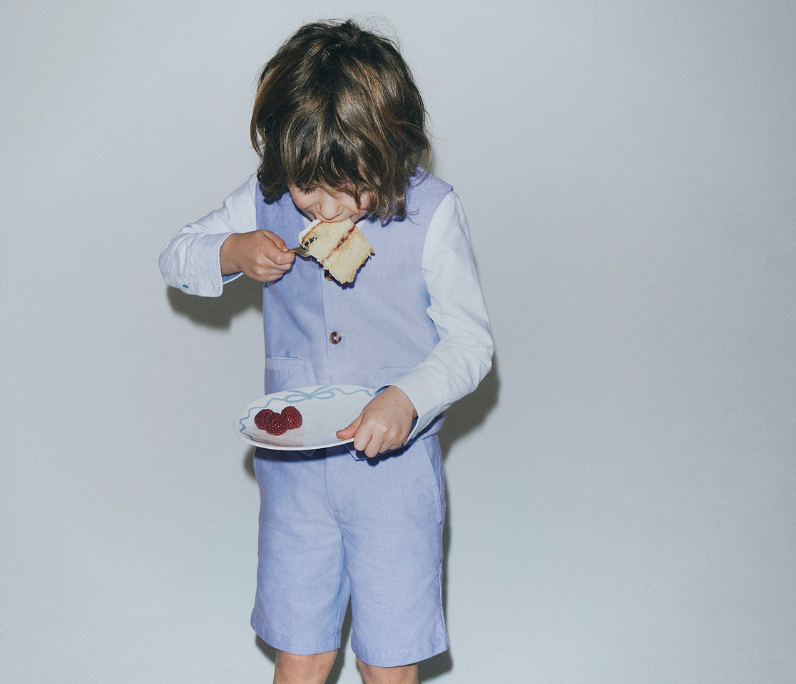 Child in a light blue outfit eating a sandwich with a white plate on a plain background