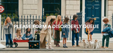 A group of children and two small dogs standing in a queue on a street in front of a building with a blue door. Large white text across the center reads, 'PLEASE FORM A DISORDERLY QUEUE.