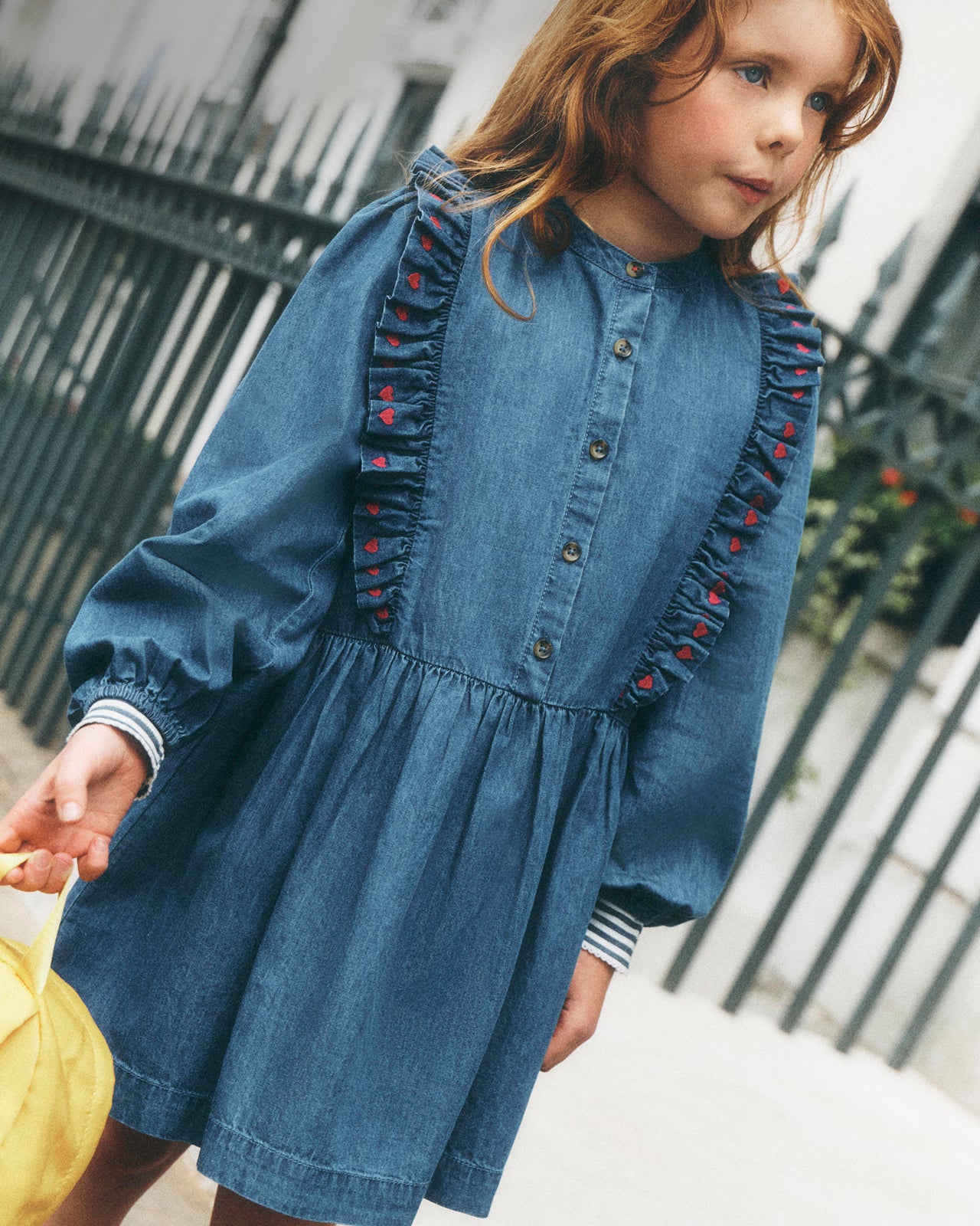 A young girl wearing a blue denim dress with ruffle details and red embroidered accents, standing outdoors in front of a dark metal fence.