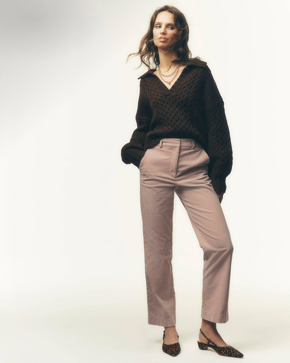 A woman modeling a dark brown textured knit sweater and light brown straight-leg trousers against a plain light gray studio background.