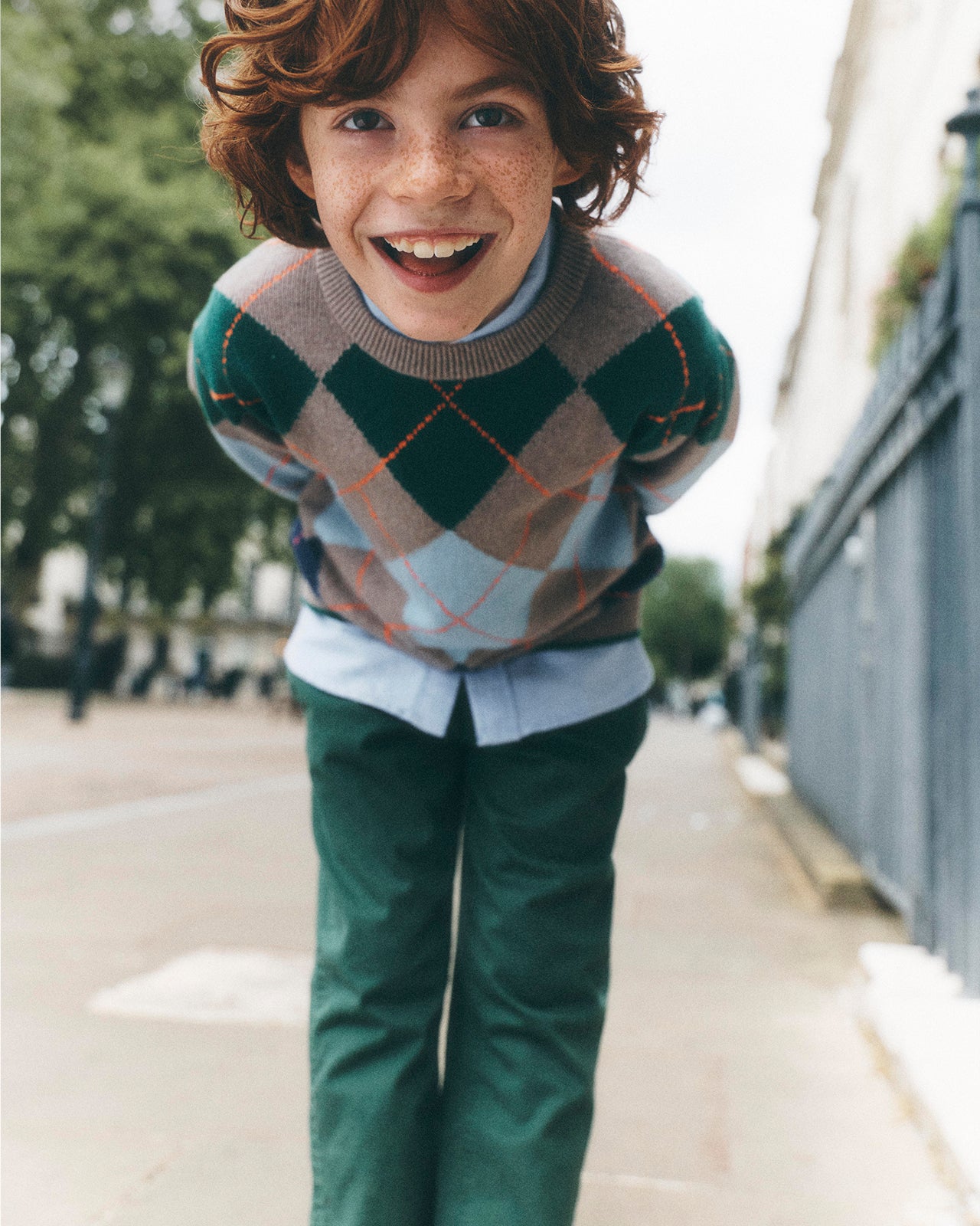 A smiling young boy with curly brown hair and freckles leans forward on a city sidewalk, wearing an argyle sweater with green, beige, and light blue diamonds, green trousers, and a light blue collared shirt.