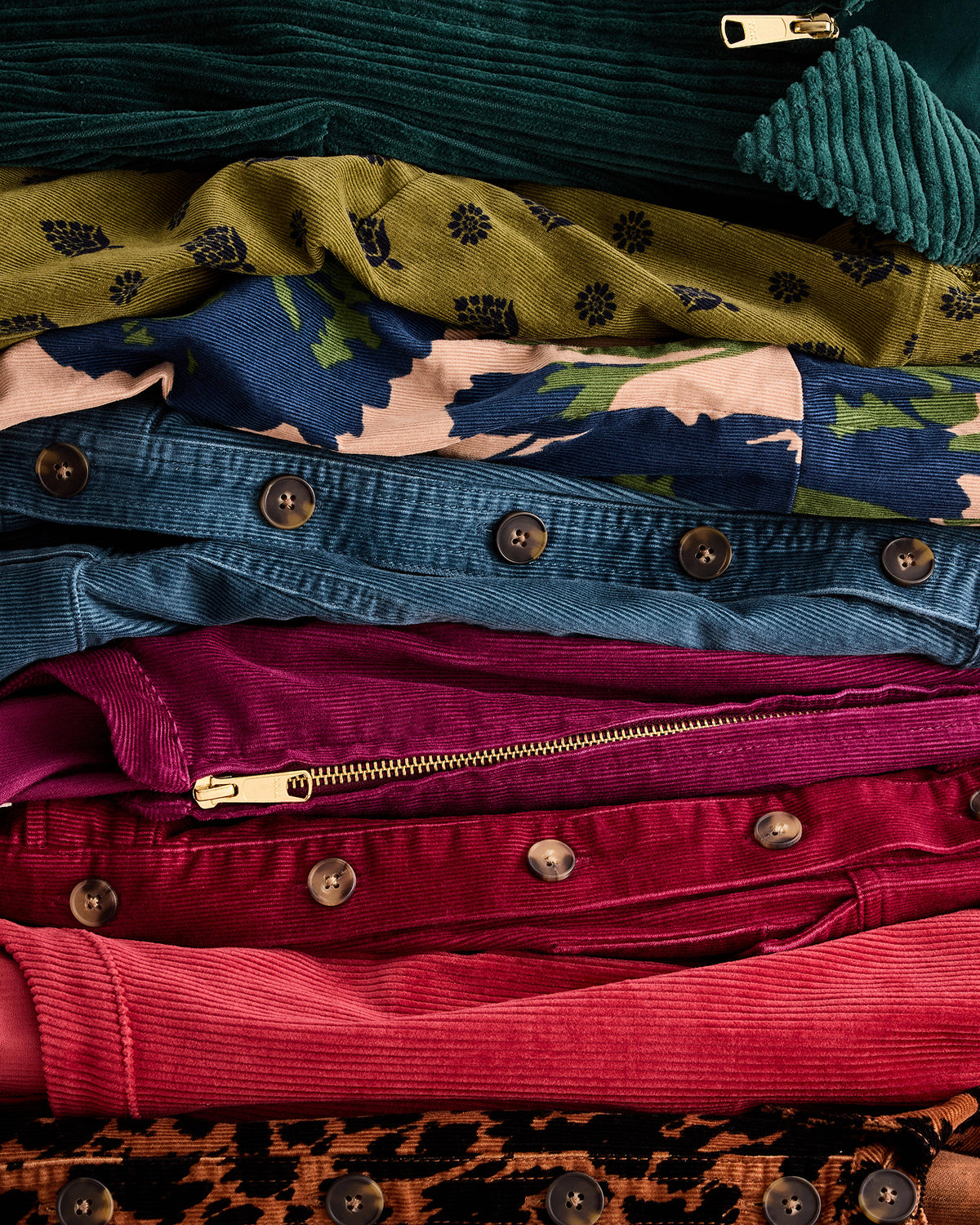 A close-up stacked arrangement of folded garments in multiple colors and textures, including teal corduroy, olive floral fabric, blue patterned shirt, denim jacket, magenta corduroy, and pink fabric, with visible metal buttons and zippers.