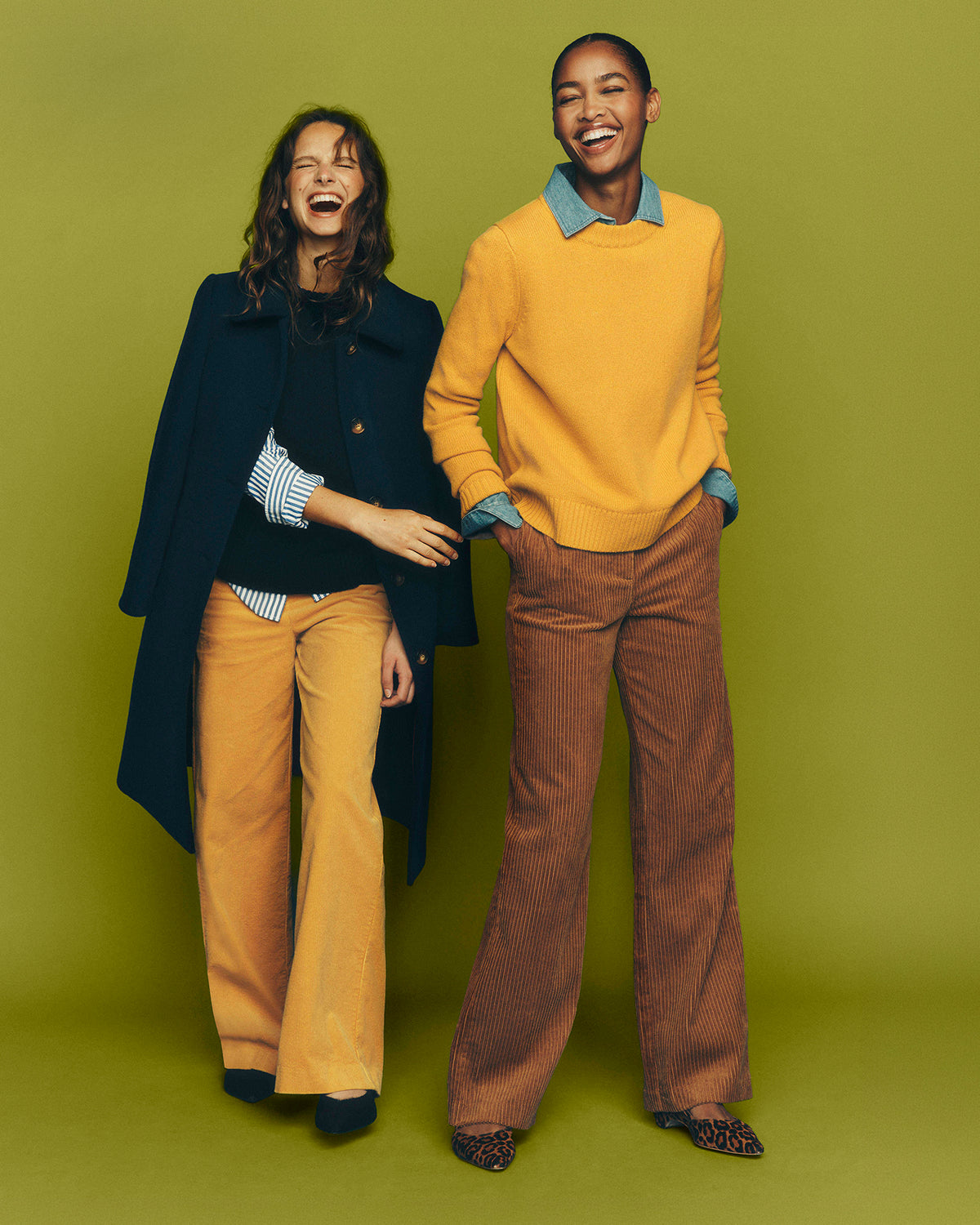 Two models posing together against a solid olive green studio backdrop. The model on the left wears a dark navy coat over a striped shirt and mustard yellow trousers.