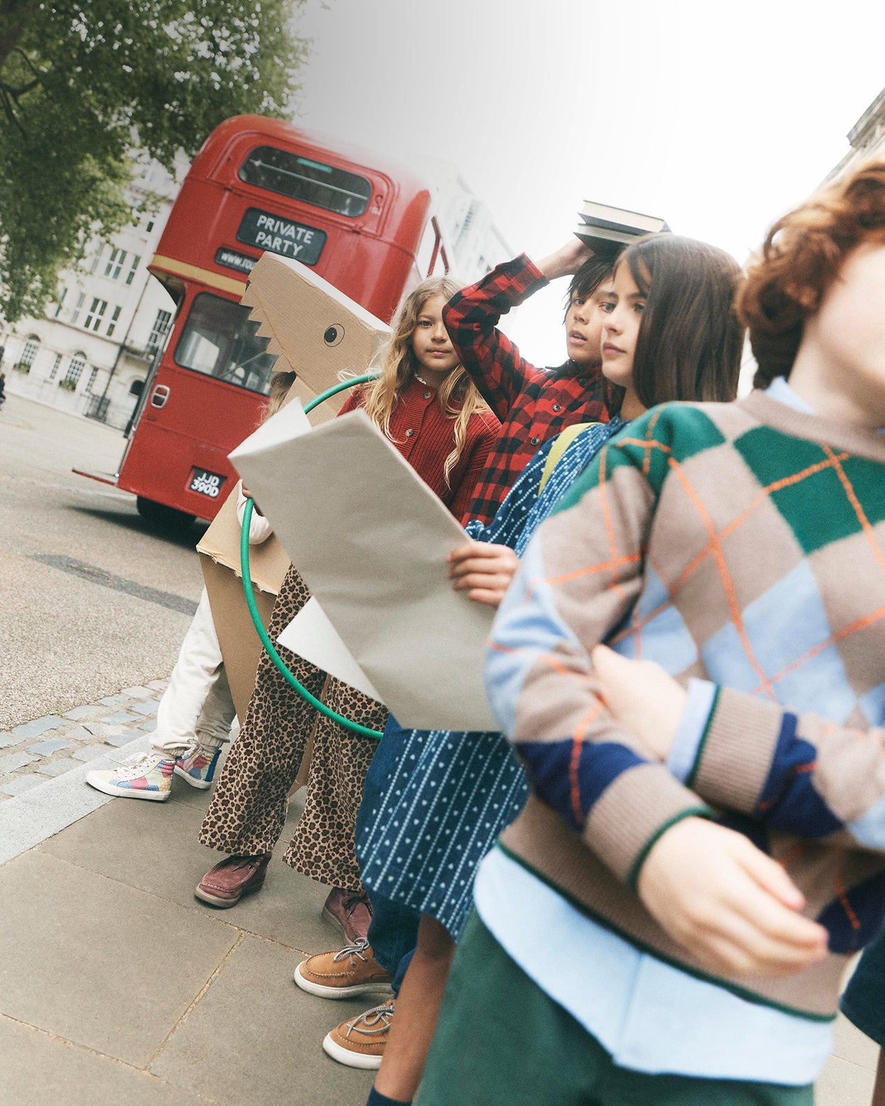 A group of children standing on a city sidewalk near a red double-decker bus, holding oversized cardboard letters that spell out a word. One child in the foreground wears a green, blue, and beige argyle sweater, and another holds a cardboard letter shaped like a dinosaur head.