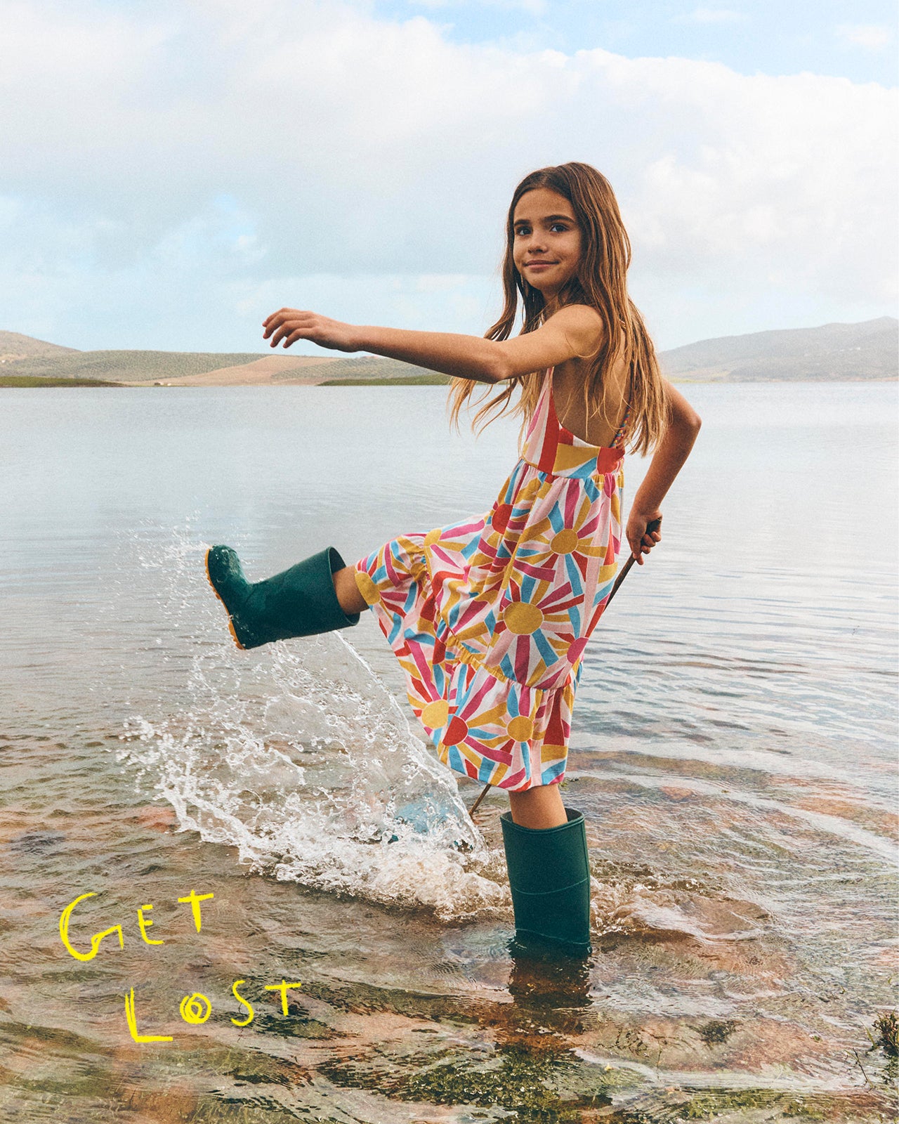 Girl in a colorful dress and green wellies playing in water with 'Get Lost' text.