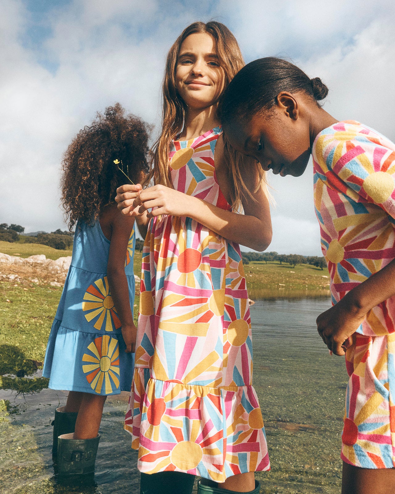 Three young girls in colorful dresses standing by a lake.