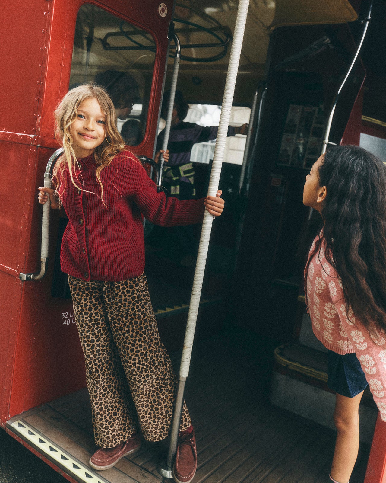 A young girl stands on the open door of a red bus, wearing a red knit cardigan and leopard-print pants, holding a grab handle. Another girl is visible in the doorway of the bus.