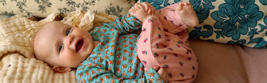 A baby lying on a couch wearing a teal long-sleeve top with a small orange and yellow pattern, paired with pink pants featuring a small floral print.