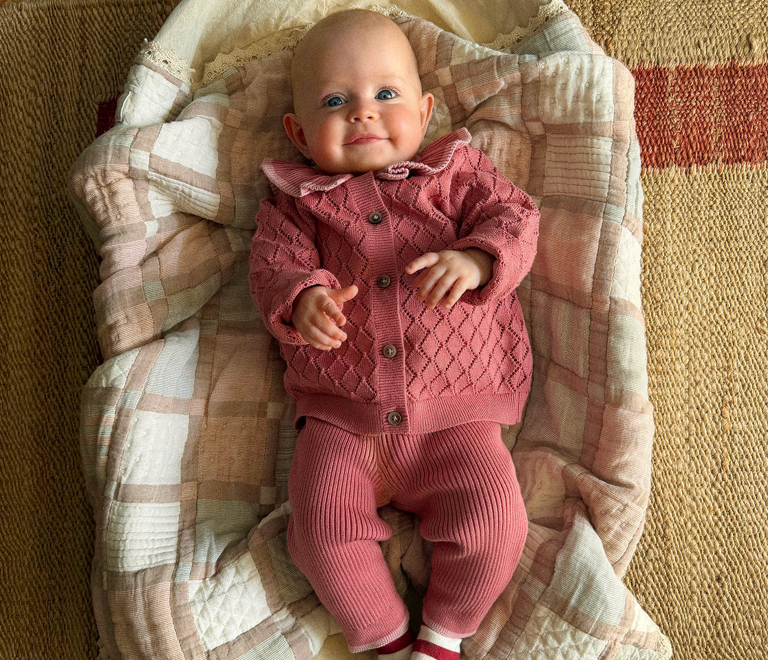 A baby lying on a quilted blanket, wearing a dusty pink knit cardigan and matching ribbed pants.