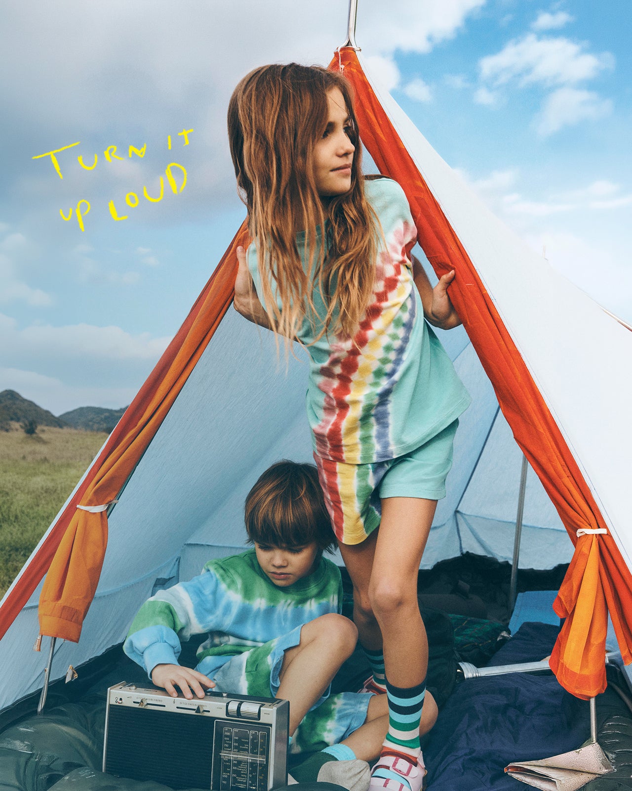 Two children in tie-dye shirts inside a tent with a clear sky and open field in the background.