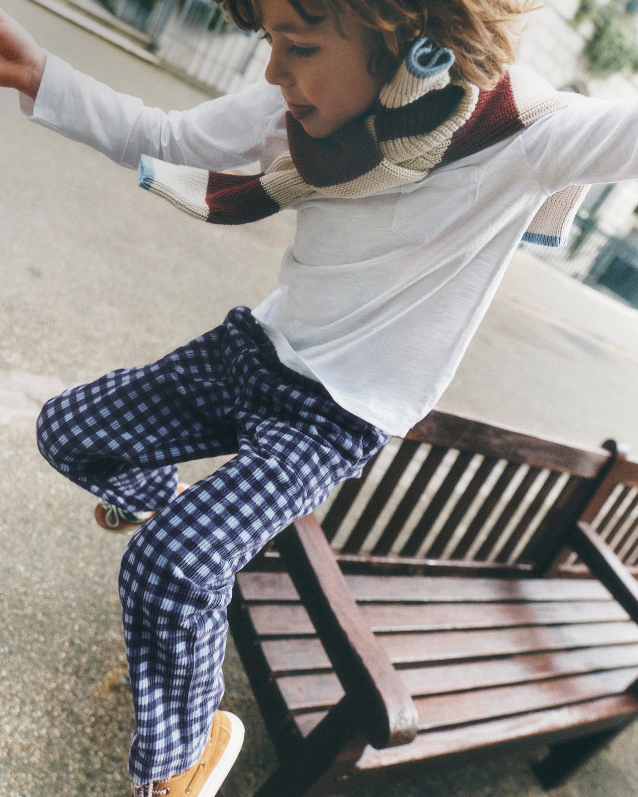 A child wearing a white long-sleeve top, a cream and burgundy knit scarf, and navy blue and white gingham pants, captured mid-motion while sitting on a wooden bench outdoors.