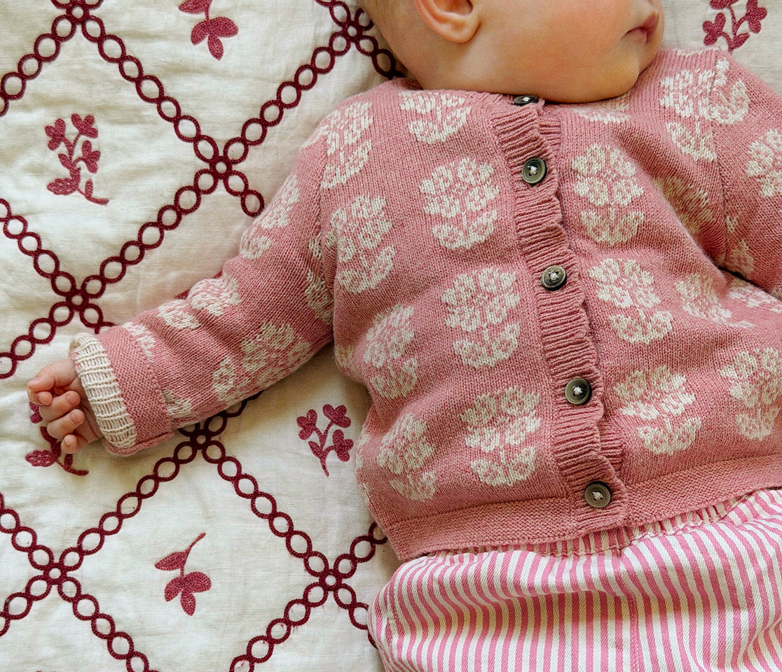A close-up of a baby wearing a pink knit cardigan with cream floral patterns and a button front, lying on a cream quilt with dark red embroidered diamond motifs.