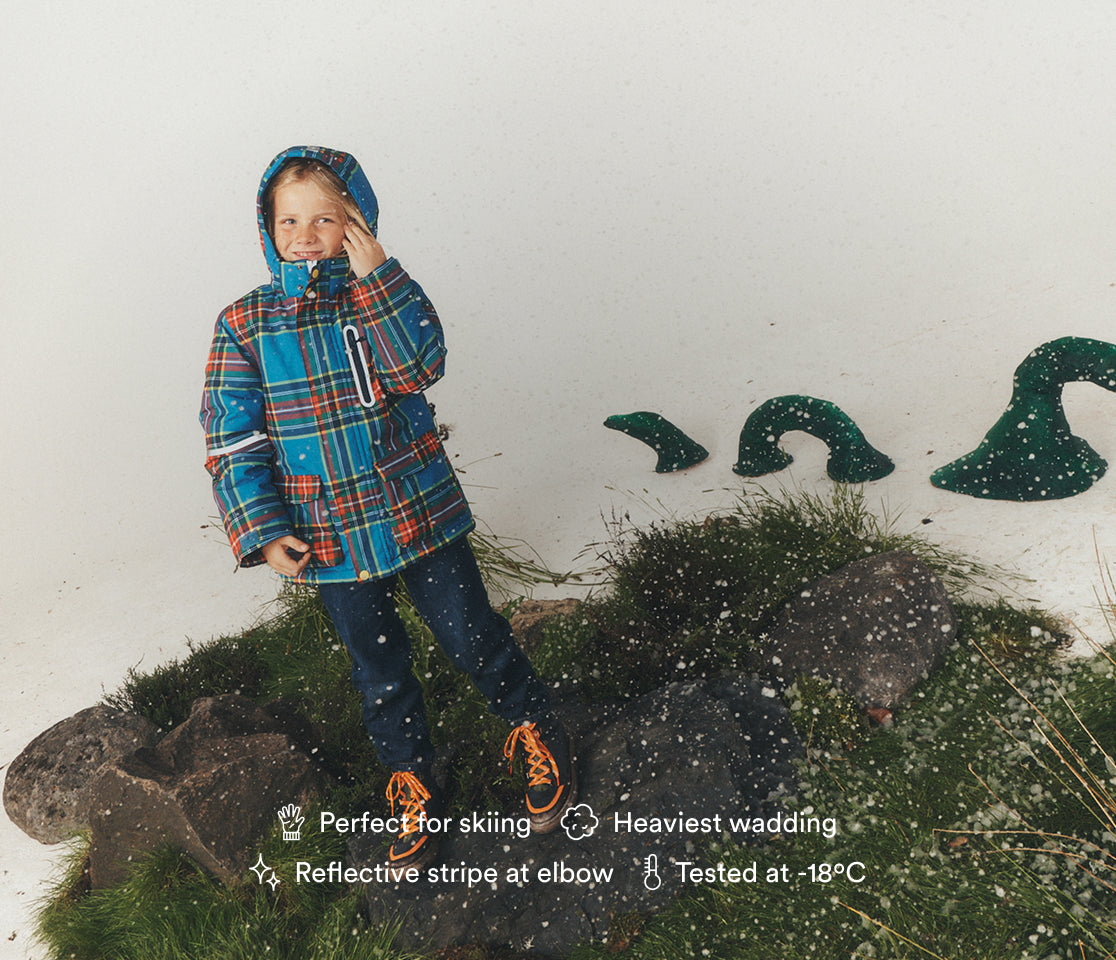 A child wearing a blue, red, and orange plaid hooded winter jacket and dark pants stands on a grassy, rocky display against a white studio background. Snowflake graphics and text call out product features like 'Perfect for skiing,' 'Heaviest wading,' 'Reflective stripe at elbow,' and 'Tested at -18°C.