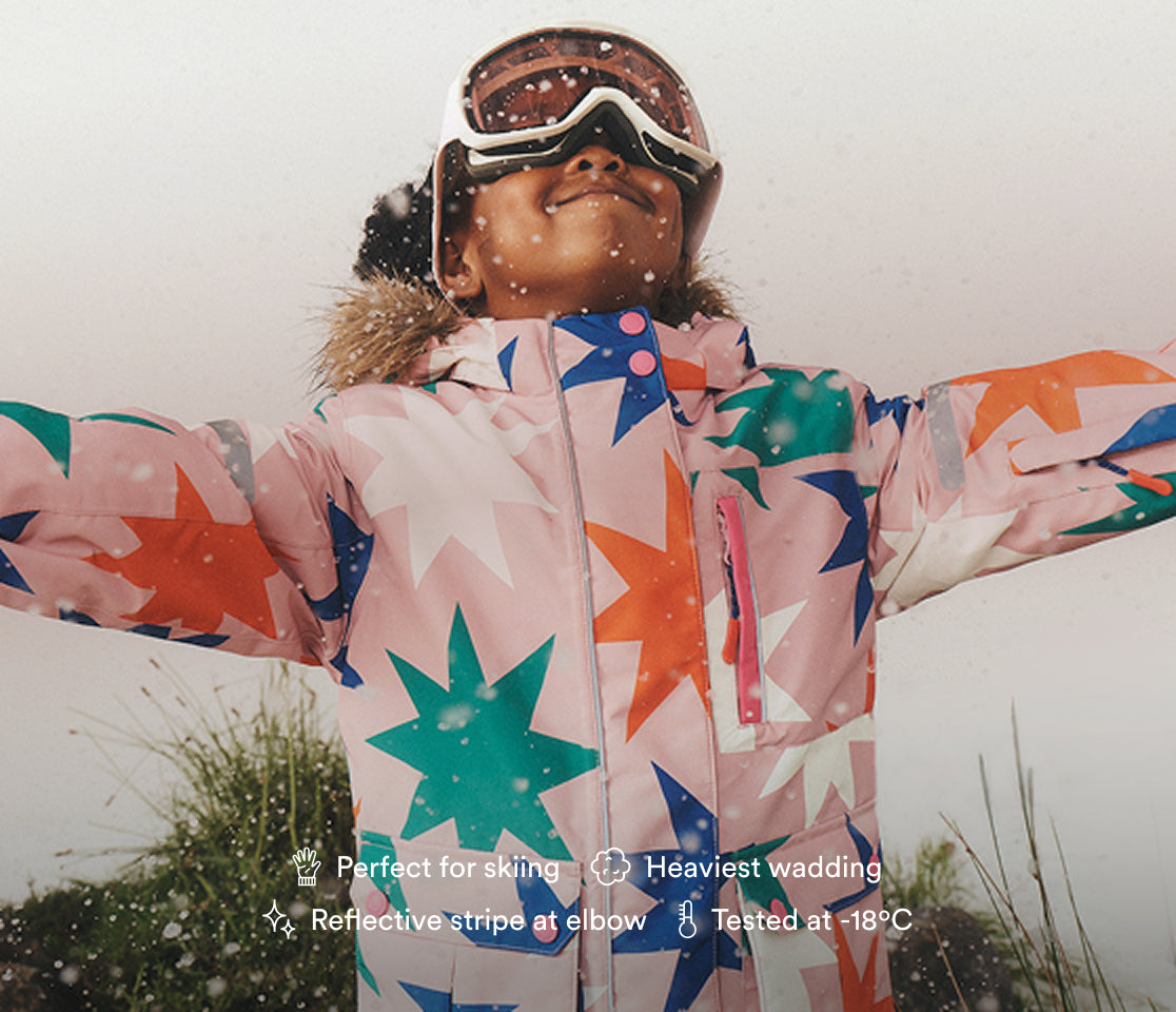 A child wearing a pink ski jacket with a colorful star pattern, goggles, and a fur-trimmed hood, standing outdoors in the snow with arms outstretched. Product text overlays are visible at the bottom.