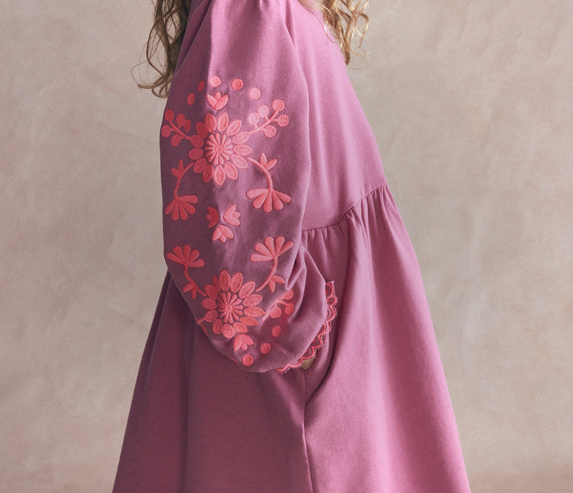 A close-up of a dusty pink dress with vibrant coral-pink floral embroidery on the sleeve, shown on a model against a neutral beige background.