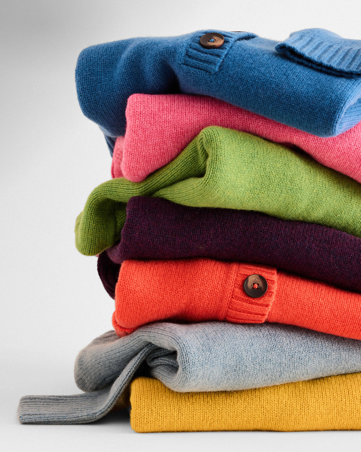 A stack of folded knit cardigans in multiple colors, including blue, pink, green, purple, orange, light gray, and mustard yellow, arranged on a light gray background.