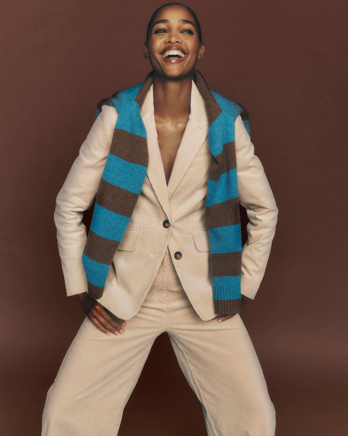 A smiling model wearing a cream-colored blazer and matching trousers, styled with a blue and brown striped knit sweater vest, posed against a solid dark brown studio background.