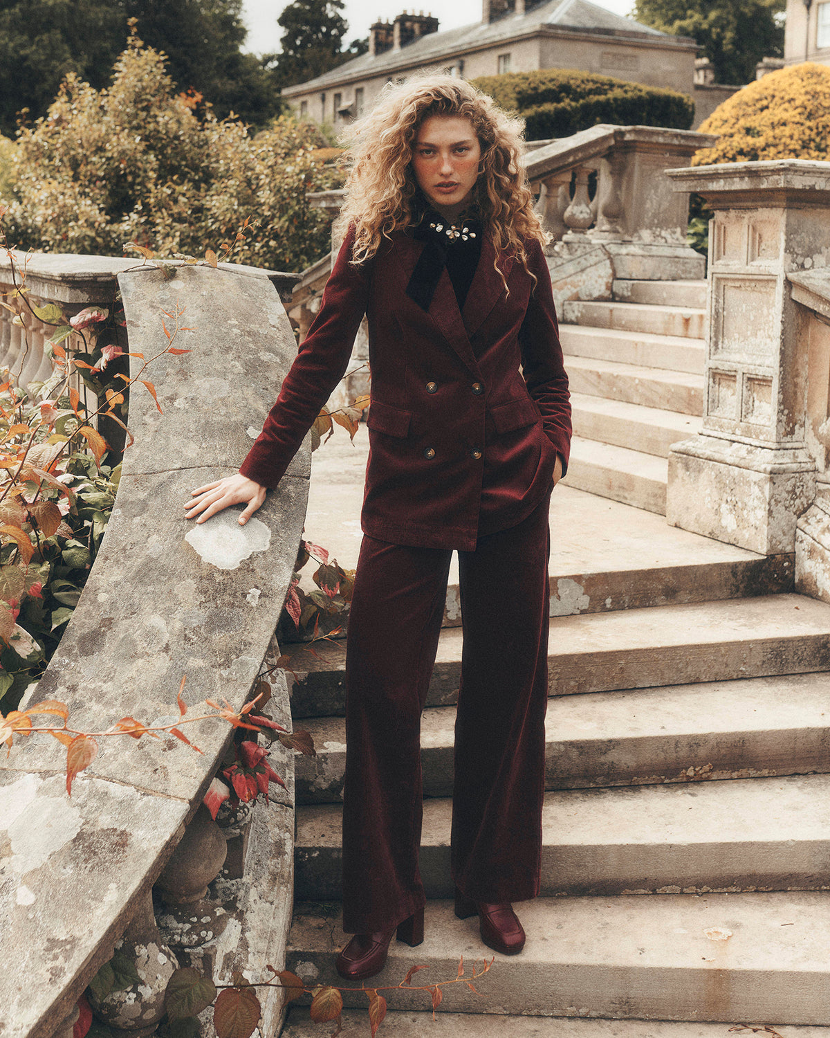 A woman stands on outdoor stone steps wearing a matching burgundy velvet blazer and wide-leg trousers suit, styled with a black high-neck top and brown leather shoes.