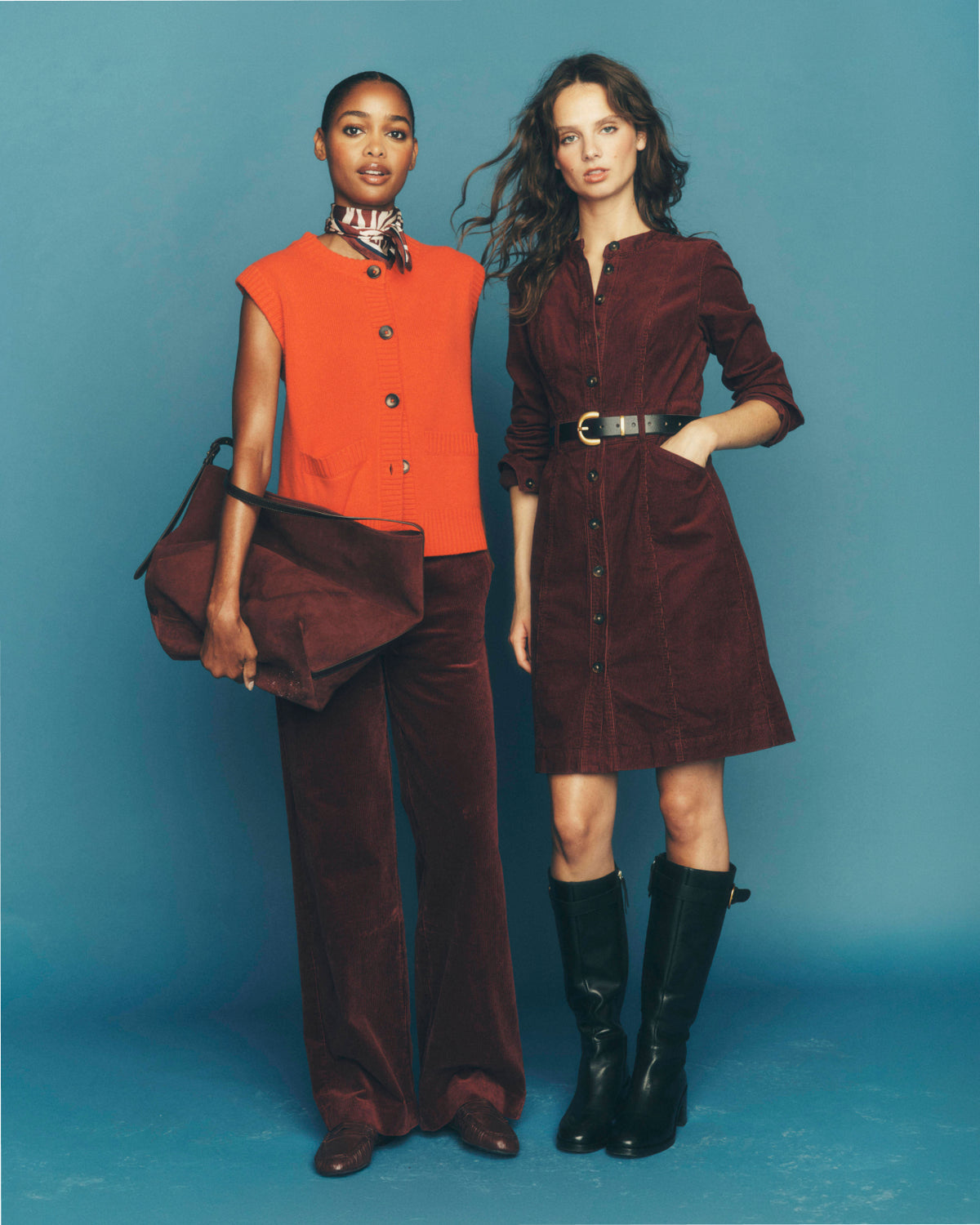 Two models posing in a studio setting against a solid blue backdrop. The model on the left wears a bright orange sleeveless top paired with dark brown velvet trousers and holds a matching dark brown bag.