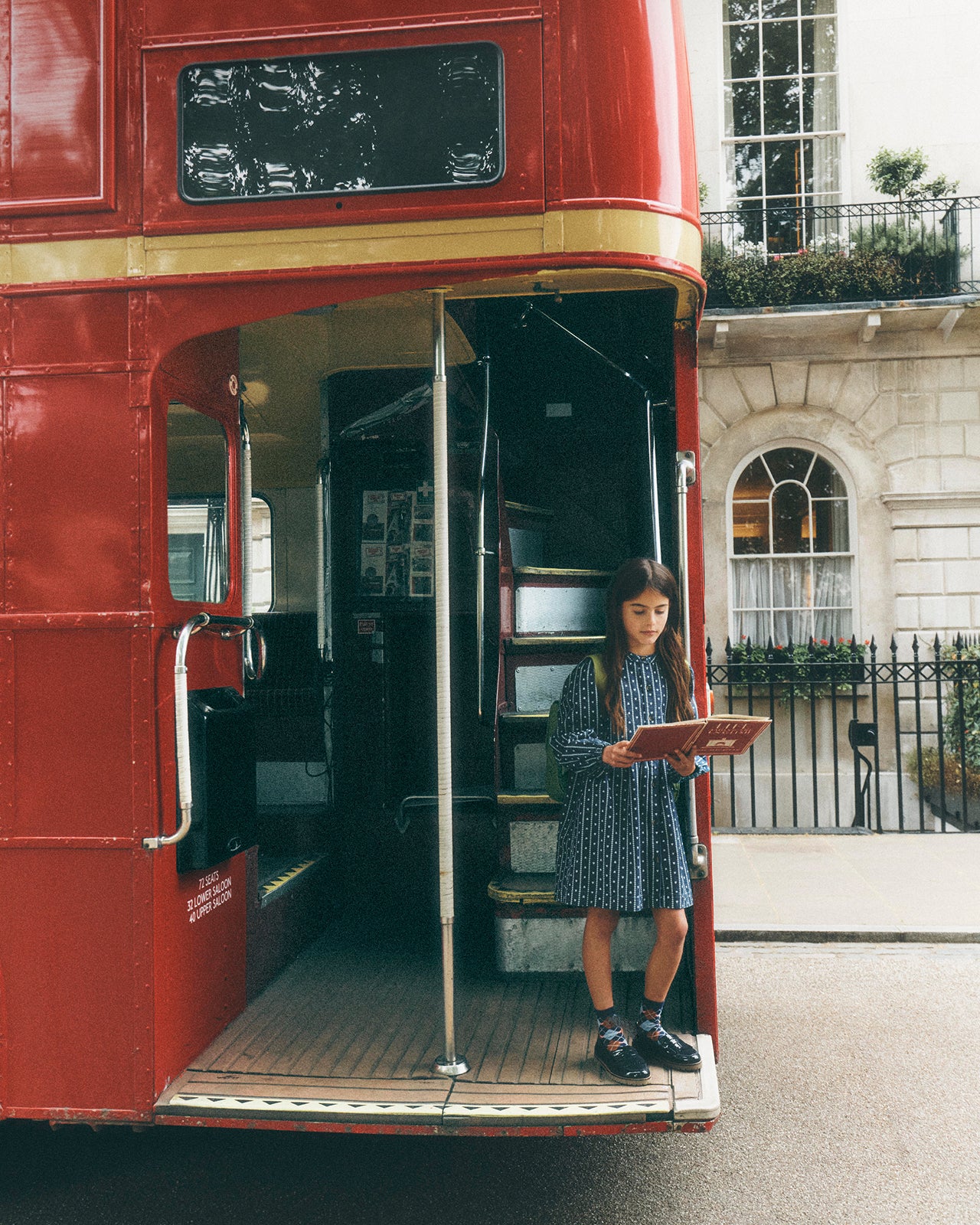 A young girl in a blue patterned dress stands at the open rear doorway of a classic red double-decker bus, holding a tablet. The photo is taken in an urban street setting with a stone building in the background.