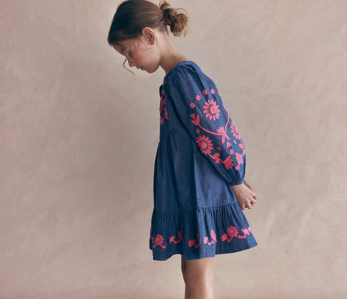 A young girl wearing a dark blue denim dress with bright pink floral embroidery, standing against a textured beige background.